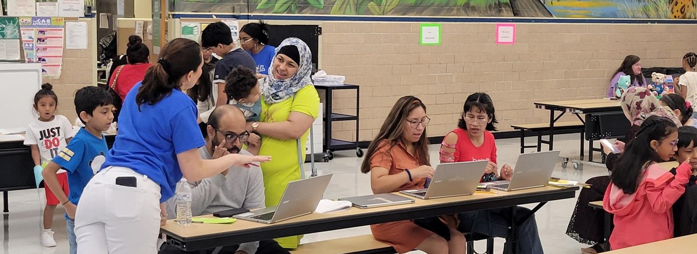 parents sitting at a cafeteria table on the computer, students and teachers standing around